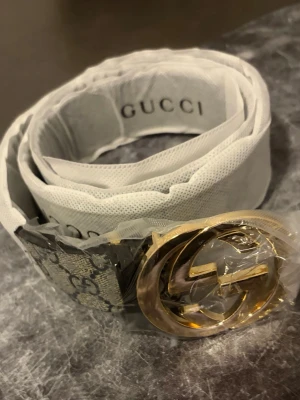 Gucci GG Supreme bälte med gulddetalj - Snyggt Gucci-bälte i beige och grått med klassiskt GG Supreme-mönster och stor guldfärgad dubbel-G-spänne. Perfekt accessoar för att lyfta vilken outfit som helst. Bältet är gjort i skinn och har exklusiv känsla.
