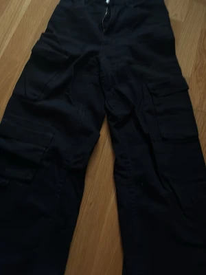 Svarta cargobyxor med fickor - Säljer ett par svarta cargobyxor med flera stora fickor på sidorna. Byxorna har en avslappnad passform och raka ben, perfekta för en streetwear-look. Tillverkade i ett slitstarkt tyg som passar till vardagsstilen.