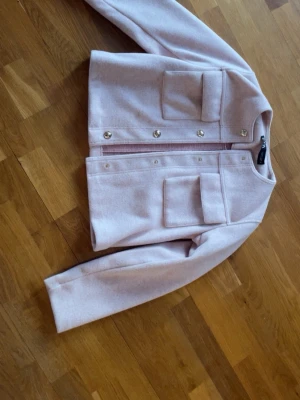 Ljusrosa overshirt med fickor - Snygg ljusrosa overshirt med raka linjer och två stora bröstfickor med lock. Jackan har guldiga knappar framtill och är gjord i ett mjukt, filtliknande material. Perfekt för lager på lager och ger en clean look till din outfit.