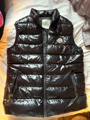 Svart dunväst från Moncler - Snygg svart dunväst från Moncler med glansig finish och klassisk logga på bröstet. Västen har två dragkedjeförsedda fickor, hög krage och quiltad design. Insidan har en unik serieteckningsdetalj. Perfekt för lager-på-lager och streetstyle.