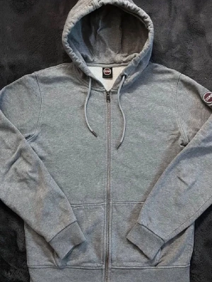 Grå Colmar hoodie med dragkedja och luva - Klassisk grå hoodie från Colmar med hel dragkedja, luva med snörning och två fickor framtill. Mjuk och bekväm i bomullsmix, perfekt för en avslappnad stil. Riktigt snygg med Colmar märket till sidan av armen perfekt för kommande vår men inte passar alla säsonger. L men passar M bra med