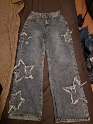 Gråa jeans med stjärnslitningar Romwe - Säljer ett par gråa raka jeans från Romwe med stora slitningar i form av stjärnor på benen. Jeansen har hög midja, fem fickor och cool tvättad look. Perfekta för dig som vill sticka ut med en unik och trendig stil.
