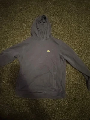 Mörkblå hoodie från Lacoste - Mörkblå hoodie från Lacoste med klassisk huva och den ikoniska lilla krokodilloggan på bröstet. Hoodien har en enkel design med ficka framtill och ribbade muddar. Perfekt för en avslappnad och stilren look.