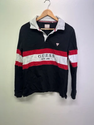 Svart långärmad topp från Guess - Svart långärmad topp från Guess med breda vita och röda ränder över bröstet och armarna. Klassisk krage i grått och vit, samt logga och texten 'Guess Est. 1981' broderat framtill. Perfekt för en sportig och avslappnad look.