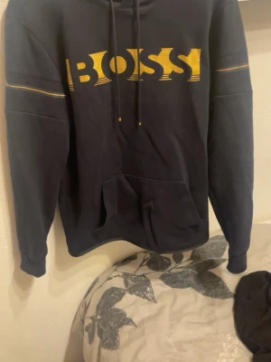 Boss hoodie svart med guldigt tryck - Svart hoodie från BOSS med stor guldig logga framtill och gula detaljer på ärmarna. Klassisk känguruficka och justerbar huva med snören. Tröjan är storlek S men passar nästan mig som vanligtvis har L