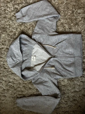 Grå zip-hoodie med huva - Säljer en ljusgrå hoodie från pull and bear. med dragkedja och huva. Tröjan har ribbade muddar och två fickor framtill. Materialet är mjukt och bekvämt, perfekt för att slänga på sig när det är lite kyligt. Klassisk och enkel design med vita snören i huvan.