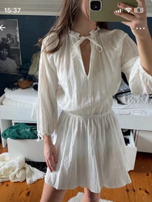 Vit kortklänning med knyt - Säljer en vit kortklänning med knyt vid halsen och v-ringad öppning. Klänningen har trekvartsärmar och är gjord i ett lätt, strukturerat bomullsmaterial. Perfekt för varma dagar och enkel att styla med accessoarer.