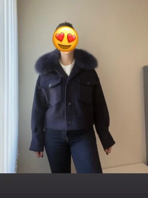 Meotine Jacka i marinblå - Snygg mörkblå kappa i kort modell med bred, fluffig pälskrage. Två stora fickor framtill med lock och knappar. Rak passform och långa ärmar med breda muddar. Perfekt för dig som vill ha en trendig och varm jacka till hösten eller vintern.