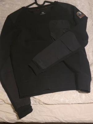 Svart sweatshirt från Parajumpers junior 14 - Cool svart sweatshirt från Parajumpers med rund hals, lång ärm och en stor ficka med dragkedja på bröstet. Patch med logga på ena ärmen och ribbade muddar. Perfekt för en streetwear-look. Oanvänd! Köpt på kidsbrandstore. Storlek 14/xs