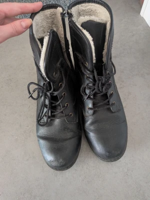 Svarta fodrade kängor med snörning - Svarta kängor i läderimitation med snörning och dragkedja på sidan. Insidan är fodrad med mjukt vitt fuskpälsfoder för extra värme. Grov sula och låg klack, perfekta för kyligare dagar. Klassisk och stilren design som passar till många outfits.