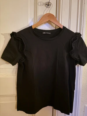 Svart volangtopp från Zara - Svart topp från Zara i mjuk bomull med korta ärmar och volangdetaljer på axlarna. Enkel rund halsringning och rak passform. Perfekt för dig som gillar stilrena plagg med en twist. Storlek M, men liten i storlek, närmare storlek S/XS. 
