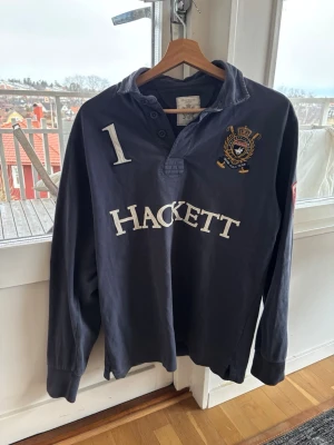 Mörkblå långärmad Hackett henley - Mörkblå långärmad henley-skjorta/rugbytröja från Hackett med broderad logga och nummerdetalj på bröst och rygg. Storlek M, mycket fint skick. Hör gärna av er vid frågor
