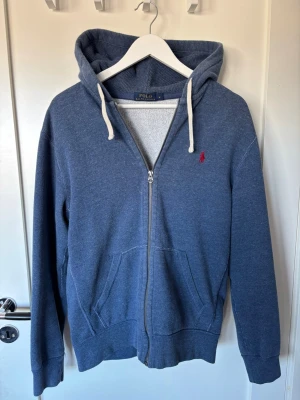 Blå zip-hoodie från Polo Ralph Lauren - Lite tjockare tvättad blå hoodie med dragkedja från Polo Ralph Lauren. Klassisk design med huva och vita snören, två fickor fram och röd broderad logga på bröstet. Perfekt inför våren då den är lite tjockare och varmare. 