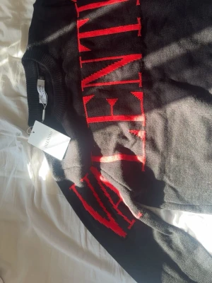 Svart stickad tröja från Valentino - Snygg svart stickad tröja från Valentino med stort rött logotyp-tryck över bröstet och ärmarna. Tröjan har rund halsringning och ribbade muddar. Perfekt för dig som gillar exklusiva och trendiga plagg med tydlig branding.