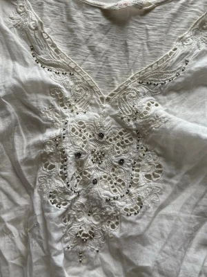 Vit blus med spets och broderi - Säljer en luftig vit blus med v-ringning och korta ärmar. Framsidan har detaljerad spets och broderade mönster med små pärlor. Storlek 1 vilket motsvarar S