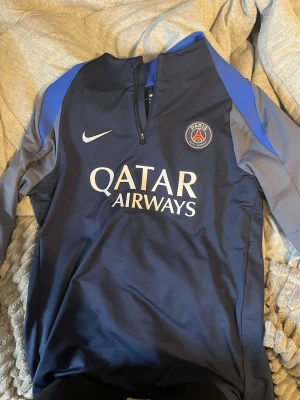 PSG träningsjacka Nike blå L - Snygg blå PSG träningsjacka från Nike med halv dragkedja och Qatar Airways-tryck på bröstet. Jackan har klubbmärke, vita och blå detaljer samt är tillverkad i Dri-Fit material för bra andningsförmåga. Slim fit-modell som passar perfekt till träning eller sport.