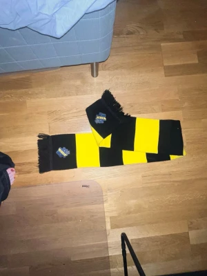 AIK halsduk svart och gul - Säljer en klassisk AIK halsduk i svart och gult med breda ränder och klubbens logga på båda sidor. Halsduken har fransar i ändarna och är perfekt för att visa ditt stöd på matchen. Materialet känns mjukt och värmande.