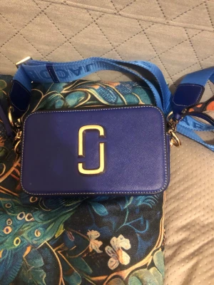 Blå Marc Jacobs Snapshot handväska - Säljer en blå Marc Jacobs Snapshot handväska i läder med bred axelrem och guldfärgade detaljer. Väskan har en rektangulär form, stort logospänne framtill och dragkedja med Marc Jacobs-graverad zip. Perfekt för dig som vill ha en statementväska.