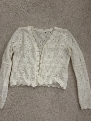 Vit stickad kofta med spetsmönster - Söt vit stickad kofta med lång ärm och vågig kant. Koftan har ett detaljerat spetsmönster med romber och hålstickning, vilket ger en luftig och feminin känsla. Perfekt att slänga över en topp för en trendig lager-på-lager-look.