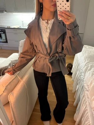 Kort grå trenchcoat med knytskärp Gina tricot  - Snygg kort trenchcoat i grått med bred krage, knytskärp i midjan och dekorativa knappar. Jackan har långa ärmar med spänne vid ärmslut och en avslappnad passform. Perfekt för dig som vill ha en trendig och stilren look.