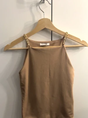 Beige linnetopp med guldkedjor.  - Snygg beige linnetopp med tunna axelband i guldfärgad kedja. Toppen har en rak halsringning.  Materialet känns mjukt och följsamt mot huden.