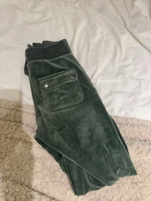 Mörkgröna mjukisbyxor Juicy Couture - Mörkgröna mjukisbyxor från Juicy Couture i mjuk velour. Byxorna har bred resår i midjan med dragsko, stora fickor fram och bak, samt en liten broderad logga på ena fickan. Perfekta för en chill och avslappnad stil.