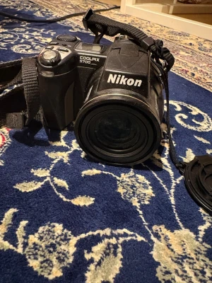 Nikon Coolpix 5700 digitalkamera - Säljer en svart Nikon Coolpix 5700 digitalkamera med rejält grepp, stor lins och tydliga knappar. Kameran har en robust design i plast och metall, samt medföljande axelrem och objektivlock. Perfekt för dig som vill ta snygga bilder med en klassisk kamera.