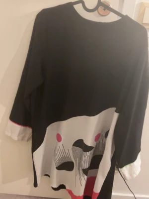 Svartvit stickad tröja med grafiskt tryck - Unik stickad tröja i svart och vitt med ett stort grafiskt ansikte framtill. Tröjan har långa ärmar och detaljer i rosa och rött på ärmslut och nederkant. Oversized passform och rund halsringning. Perfekt för dig som gillar statement-plagg.