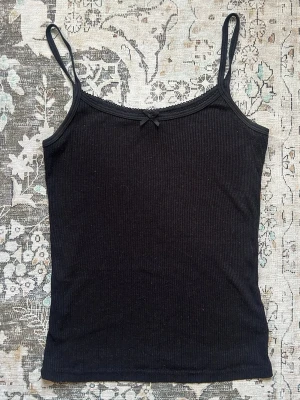 Svart ribbad linnetopp med rosett - Svart ribbad linnetopp med smala axelband och en liten rosett framtill. Toppen har en enkel och stilren design som passar perfekt till jeans eller kjol. Materialet är mjukt och stretchigt för en bekväm passform.