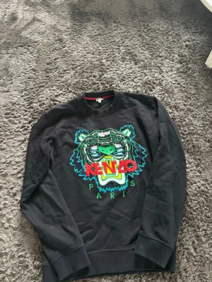 Svart Kenzo sweatshirt med tiger - Cool svart sweatshirt från Kenzo med det ikoniska tigerhuvudet broderat i neonfärger på bröstet. Tröjan har rund halsringning, ribbade muddar och detaljer i blått, grönt, rött och gult. Perfekt statement-plagg för dig som gillar streetwear.