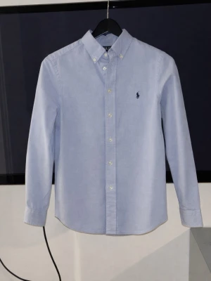 Ljusblå skjorta från Ralph Lauren - Klassisk ljusblå skjorta från Ralph Lauren med button-down krage och broderad logga på bröstet. Skjortan har långa ärmar och knäppning framtill. Storlek 14 åring. Använd lite men inte mycket