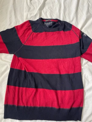 Randig stickad tröja Nautica Jeans XL - Snygg stickad tröja från Nautica Jeans Co. i storlek XL. Tröjan har breda marinblå och röda ränder, rund halsringning och ribbade muddar. Perfekt för dig som gillar klassisk streetwear med en sportig touch.