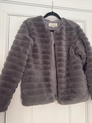 Grå pälsjacka från Pomed Lux - Säljer en fluffig grå pälsjacka från Pomed Lux med snygga horisontella ränder och guldfärgad dragkedja. Jackan är kortare i modellen och har långärmade ärmar. Insidan är fodrad med diskret prickigt tyg. Perfekt för dig som gillar mjuka och trendiga jackor.