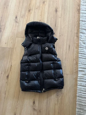 Svart dunväst från Moncler med huva - Snygg svart dunväst från Moncler med avtagbar huva och två dragkedjefickor. Västen har en glansig finish och klassisk Moncler-logga på bröstet. Fodrad med dun för extra värme och komfort. Storlek 2 vilket motsvarar M. 