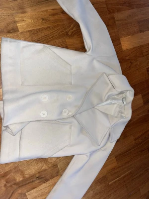 Creme vit kort kappa  - Stilren vit kappa med dubbelknäppning och stora framfickor. Kort modell med bred krage och fyra vita knappar framtill. Perfekt för dig som gillar minimalistisk och clean stil. Materialet känns mjukt och har en ull-liknande finish.
