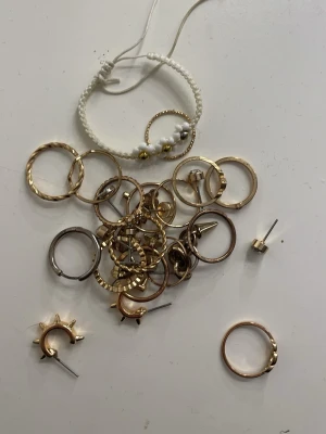 Mixade ringar och örhängen i metall - Säljer ett blandat set med ringar och örhängen i guld- och silverfärgad metall samt ett vitt armband med små pärlor och gulddetaljer. Ringarna har olika former och mönster, vissa örhängen har nitar och andra är enkla hoops. Perfekt för dig som gillar att mixa smycken.