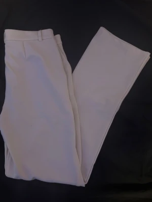 Vida kostymbyxor från 157, XS - Snygga, vida kostymbyxor i ljusbeige från lager. Byxorna har klassisk passform med hällor för skärp och stängs med dold knapp och dragkedja. Perfekta för en clean och stilren look. Materialet känns mjukt och följsamt.