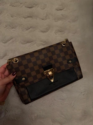 Brun-rutig handväska med guldkedja - Snygg handväska i brunt rutigt mönster med svarta detaljer och guldfärgat lås. Väskan har både kedjerem och svart axelband, samt en elegant guldspänne framtill. Perfekt accessoar för att lyfta din outfit.