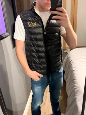 Svart dunväst från EA7 Emporio Armani - Snygg svart dunväst från EA7 Emporio Armani med guldfärgad logga på bröstet. Västen har quiltad design, dragkedja framtill och stårkrage. Perfekt att slänga över en hoodie eller t-shirt för en cool och sportig look.