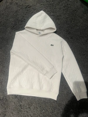 Vit kabelstickad hoodie från Lacoste - Säljer en clean vit hoodie från Lacoste med kabelstickat mönster och klassisk krokodil-logga på bröstet. Hoodien har ribbade muddar och huva, perfekt för en chill och stilren look. Materialet känns mjukt och skönt mot huden.