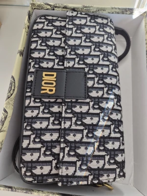 Svartvit axelväska  - Snygg axelväska fråni svartvitt jacquardtyg. Väskan har två band i svart läder och guldfärgade metalldetaljer. Magnetlås framtill och rymligt innerfack. Perfekt för dig som gillar lyxig och trendig design.