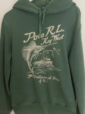  Polo Ralph Lauren Key West hoodie - Grön hoodie från Polo Ralph Lauren med tryck 'Polo RL Key West' och motiv av en fisk och båt. Klassisk huva med dragsko och ribbade muddar. Perfekt för en avslappnad stil och skön att bära. Mjuk bomullskvalitet. Nypris 2900. Riktigt bra skick. Storlek s, och nästan omöjligt att få tag på.