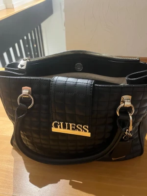 Svart quiltad handväska från Guess - Snygg svart handväska från Guess med quiltat mönster och silvriga metalldetaljer. Väskan har dubbla handtag, dragkedja upptill och en tydlig Guess-logga framtill. Perfekt för dig som vill ha en stilren och trendig accessoar. Äkta 