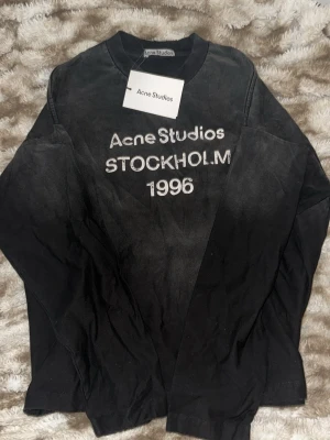Svart långärmad topp Acne Studios - Svart långärmad topp från Acne Studios med tryck framtill där det står 'Acne Studios STOCKHOLM 1996'. Rund halsringning och rak passform. Tillverkad i mjukt material som känns skönt mot huden.