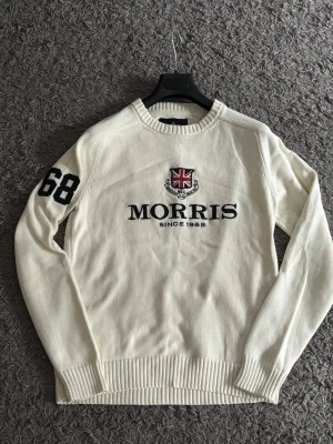 Vit stickad tröja från Morris - Snygg vit stickad tröja från Morris med broderad logga och Union Jack på bröstet samt texten 'Morris Since 1968'. Svart siffra 68 på ena ärmen. Klassisk rund halsringning och ribbade muddar. Perfekt för en stilren och sportig look.