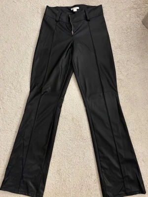 Svarta läderbyxor från Gina  - Snygga svarta läder byxor med utsvängda ben och mid midja. Byxorna har pressveck framtill, bälteshällor och dragkedja. Perfekta för en trendig och edgy look.