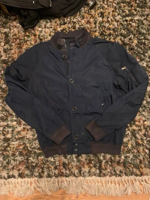 Mörkblå bomberjacka från Polo Ralph Lauren  - Säljer en riktigt snygg mörkblå bomberjacka från Polo Ralph Lauren, med bruna ribbade muddar och krage. Jackan har både dragkedja och knappar framtill samt en ficka med dragkedja på ärmen. Liten defekt (kolla bild 5) men int esp synligt. Vid funderingar, fråga på! 