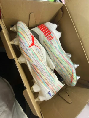 Puma Ultra färgglada fotbollsskor - STORLEK 40. Säljer ett par snygga fotbollsskor från Puma med färgglada ränder i vitt, blått, gult och rött. Metall dobbar och den klassiska röda Puma-loggan på sidan. Perfekta för dig som vill sticka ut på planen! Jag vann de på 3Arena och har signaturen av Hammarbys dam A lags (på nåt sätt utsuddat)lagkapten Alice Carlson. NYA‼️OANVÄNDA ‼️‼️