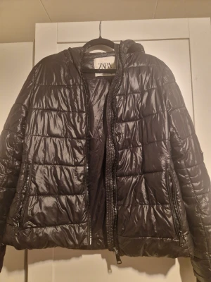 Svart pufferjacka från ZARA - Svart pufferjacka från ZARA med glansig finish och quiltad design. Jackan har huva, dragkedja framtill och två fickor med dragkedja. Perfekt för kalla vinterdagar och ger en cool streetstil. Köpte den för 800 säljer för 500. Använda 1-2 gånger?