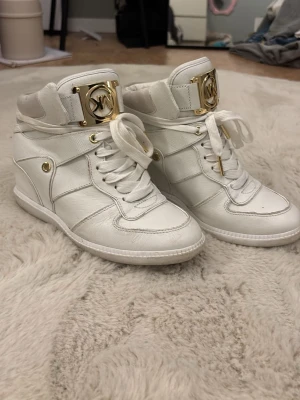Vita high top sneakers från Michael Kors (går it att köpa längre) - Snygga vita high top sneakers från Michael Kors med guldfärgade detaljer och stort MK-spänne på plösen. Skorna har vita snören, guldfärgade nitar och är tillverkade i ett strukturerat material som ger en exklusiv känsla. Perfekta för dig som vill sticka ut.